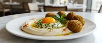 Creamy hummus topped with a fried egg, fresh herbs, olive oil, and crispy falafel balls served on a white plate in a restaurant.