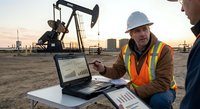 Oil field engineer in safety vest analyzing production data on laptop computer at drilling site during sunset with pumpjack equipment in background.