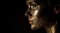 Profile of woman with gold body paint and dramatic lighting against black background, showcasing luxury cosmetics and high fashion makeup artistry.