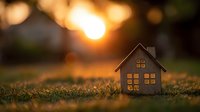 Miniature wooden house model on grass at sunset with warm golden bokeh background, symbolizing real estate, home ownership, and property investment.