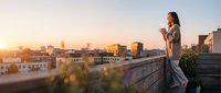 Young woman with long brown hair, Caucasian ethnicity, standing on urban rooftop at golden hour sunset, holding camera, photographing cityscape. Suitable for travel, photography, lifestyle content.