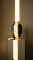 Metallic gold vase with textured surface displayed on stone pedestal, illuminated by natural light beam, creating dramatic shadows and highlights for luxury home decor.