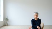 Mature woman with short gray hair, Caucasian ethnicity, wearing dark navy blazer over blue top, seated in modern minimalist office or home workspace with white walls and natural light from window, professional portrait for corporate business use.