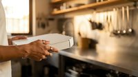 Person holding white box in kitchen with stainless steel appliances and tools on wall. Male hand presenting takeout or delivery package in modern cooking space.