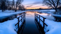 Wooden footbridge over frozen lake at sunset creates mirror reflection in water, surrounded by snow-covered landscape and bare winter trees in twilight.