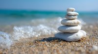 Balanced white stones stacked on sandy beach with turquoise ocean waves and foam in background, creating zen and peaceful atmosphere for wellness and spa designs.