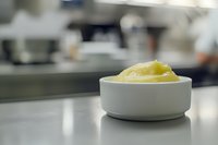 Fresh butter curls in white ceramic ramekin on stainless steel counter, soft focus background shows professional kitchen setting. Perfect for culinary content.
