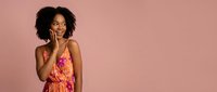 Young African American woman with curly hair showing surprised expression with hand on cheek wearing colorful floral dress on pink background.
