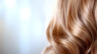 Luxurious wavy blonde hair with soft highlights and natural shine on light background, close-up view showing professional hairstyling and healthy hair texture.