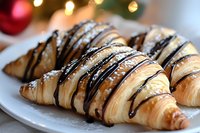 Fresh baked croissants drizzled with dark chocolate and powdered sugar on white plate, festive holiday lights and red ornament in soft focus background.