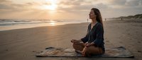 Young woman practicing meditation on beach blanket during golden hour sunset with peaceful ocean waves and sandy shore creating serene wellness atmosphere.