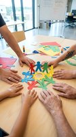 Diverse team of men and women with varying skin tones and ages collaborating around wooden table, holding colorful paper cutout figures and shapes in modern office with whiteboard and city view.