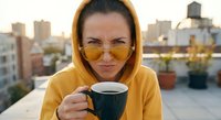 Young woman in yellow hoodie and sunglasses enjoying morning coffee on urban rooftop terrace with city skyline background during golden hour sunrise.