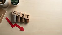 Cost reduction concept with wooden blocks spelling COST over stacked coins and red downward arrow on wooden desk for business finance planning.