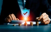 Businessman protecting financial assets with security shield symbol over coins and tablet representing investment protection and risk management.