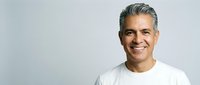 Confident middle-aged Hispanic man with gray hair smiling warmly in white casual shirt against neutral background for business and lifestyle content.