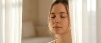 Young Caucasian woman with closed eyes enjoying peaceful moment in soft natural sunlight streaming through window curtains for wellness and mindfulness concepts.