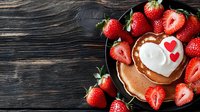 Fresh strawberries around heart-shaped pancake with whipped cream and red hearts on dark wooden background. Romantic breakfast concept for Valentine's Day.