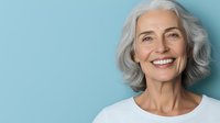 Confident mature woman with silver gray hair and genuine smile wearing white top against blue background, representing vitality and positive aging.