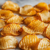 Crispy ridged potato chips with seasoning and spices scattered on gray surface, macro food photography showing detailed texture and golden color.