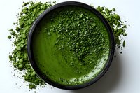 Vibrant green matcha powder in black bowl with scattered tea dust around edges on white background for culinary and wellness content.