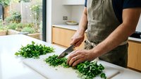 Male chef in apron chopping fresh green herbs on white cutting board in modern kitchen with garden view for culinary cooking preparation.