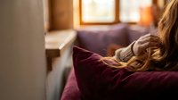 Young woman with long wavy hair sitting on a cozy sofa, gazing out a warm sunlit window in a rustic interior setting.