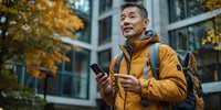 Middle-aged Asian man in yellow winter jacket using smartphone near office building with autumn foliage, expressing positive emotion while checking mobile device.