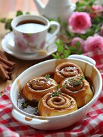 Fresh cinnamon rolls with caramel sauce in ceramic baking dish, served with coffee cup and pink roses on red checkered tablecloth, cozy breakfast setting.