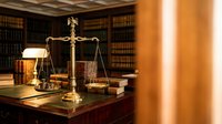 Golden scales of justice on wooden desk in traditional law library with leather-bound books creating professional legal atmosphere for attorney services.