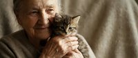 Senior woman gently holding adorable tabby kitten close to her chest, showing tender bond between elderly person and young cat for pet adoption campaigns.