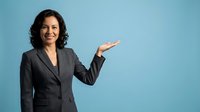 Professional Hispanic businesswoman in dark suit presenting with open palm gesture on blue background for corporate marketing materials.