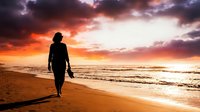 Silhouetted surfer holding board walking along sandy beach at dramatic sunset with vibrant orange and purple sky, ocean waves, and golden light reflecting on water for travel and lifestyle content.