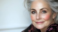 Elegant mature Caucasian woman with silver hair and natural makeup smiling confidently against light background, showcasing graceful aging and beauty in senior years.