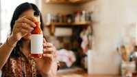 Young woman of South Asian descent holding up a blank white cosmetic bottle with orange cap in a home studio setting, suitable for beauty product marketing and skincare branding.