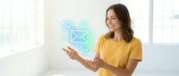 Young woman with medium skin tone wearing yellow t-shirt gestures toward glowing email icons in bright office space, representing digital communication, messaging services, and online correspondence for business applications.