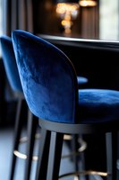 Luxurious navy blue velvet bar stool with curved backrest against blurred restaurant background, modern interior design with soft bokeh lighting effects.