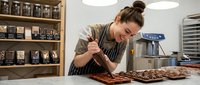 Young female artisan with brown hair in bun, wearing apron and glasses, carefully decorating chocolate confections in professional kitchen workspace with shelving and equipment.