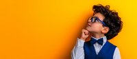 Thoughtful young Hispanic boy wearing blue vest, bow tie and glasses against vibrant yellow wall, looking up with hand on chin in contemplative pose.