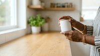 Cropped view of woman holding takeout coffee cup in modern kitchen with wooden countertop and potted plant on shelf.