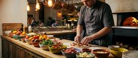 Professional chef preparing fresh ingredients in modern restaurant kitchen with wood-fired oven and warm ambient lighting for culinary service.