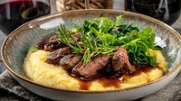Juicy medium-rare beef steak slices served on creamy polenta with sautéed greens and rosemary garnish in rustic ceramic bowl with red wine sauce.