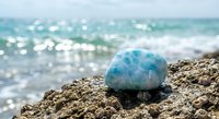 Blue larimar gemstone resting on sandy beach with turquoise ocean waves and sparkling sunlight creating peaceful coastal meditation scene.