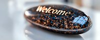 Decorative black stone with golden Welcome text and blue window symbol, covered in glitter effect against blurred background. Spiritual healing concept.