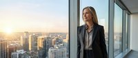 Middle-aged Caucasian woman in professional business attire standing in modern high-rise office overlooking cityscape at sunset, suitable for corporate, leadership, and business concepts.