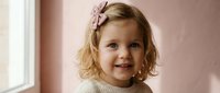 Adorable blonde toddler girl with curly hair and pink bow smiling joyfully by window in soft natural light. Perfect for family lifestyle and childhood concepts.