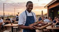Smiling African American chef in apron holding grilled meat platter at outdoor restaurant during golden hour sunset with string lights and dining guests.