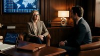 Mature woman wearing glasses and beige blazer sits at wooden desk across from man in dark suit during business meeting in modern office with world map display and financial charts on screens.