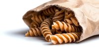 Whole grain fusilli pasta spilling from brown paper bag on white background, showcasing nutritious carbohydrate food for healthy cooking.