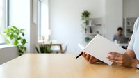 Young woman with medium skin tone in professional white shirt reading document at wooden desk in bright modern office space with plants and colleagues in background.